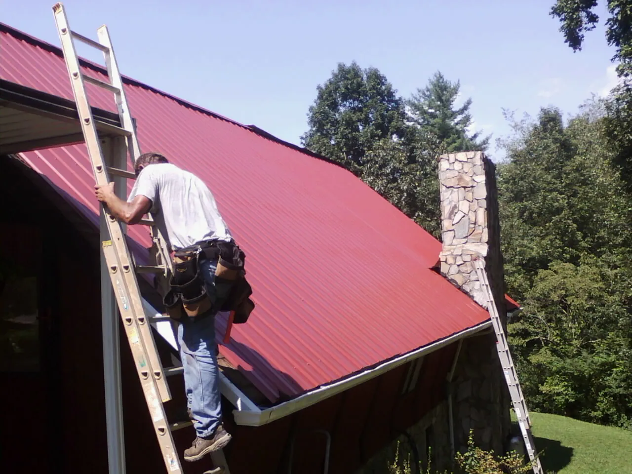 Roofer on a ladder beside a red metal roof and stone chimney — Soffit & Fascia Repair by Roofing Brothers in Eden Prairie